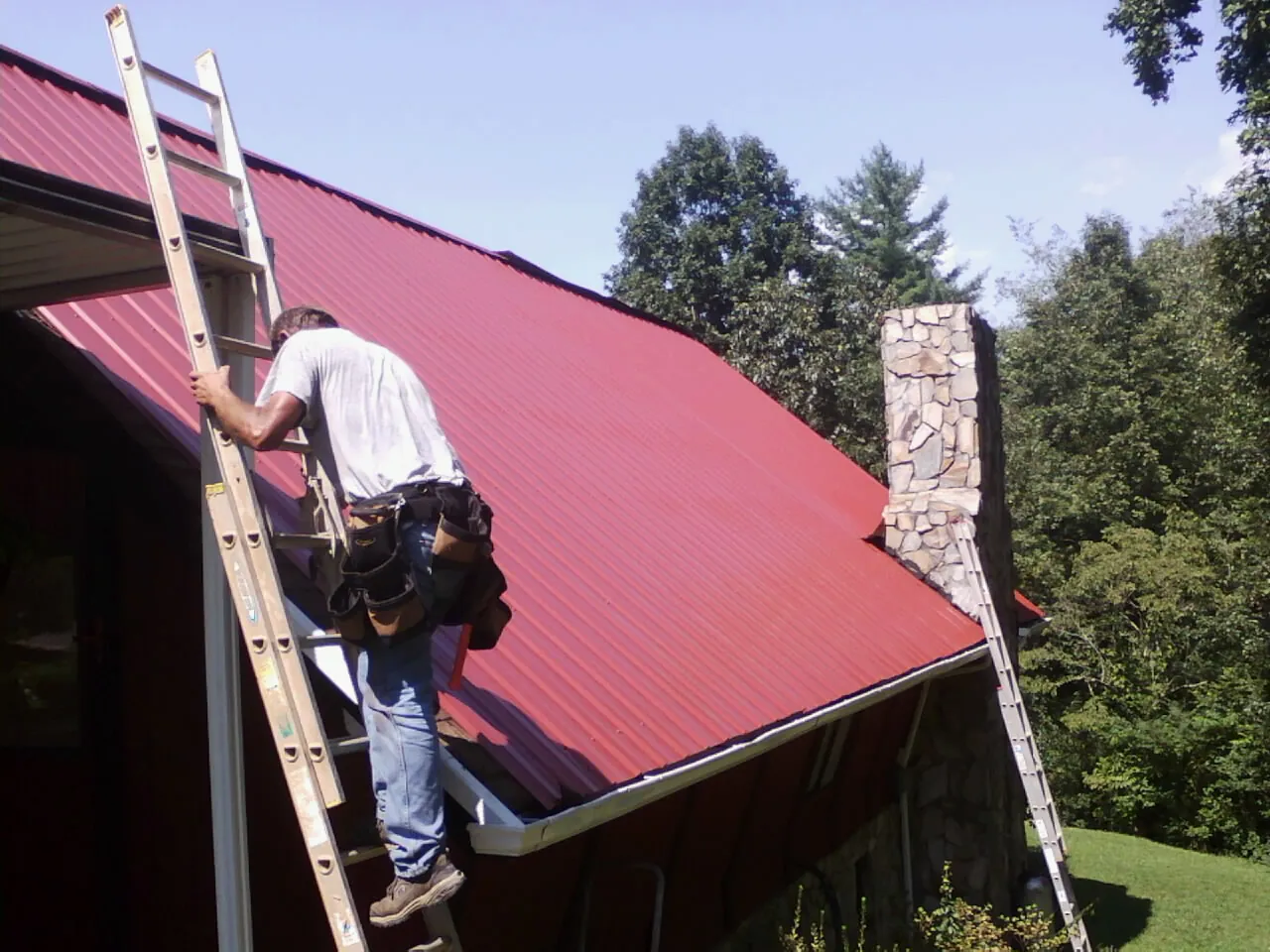 Roofer on a ladder beside a red metal roof and stone chimney — Soffit & Fascia Repair by Roofing Brothers in Arock
